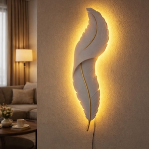 May include: A white feather-shaped wall light fixture with a gold accent. The light is illuminated from behind, creating a warm glow. The fixture is mounted on a neutral-colored wall, and a power cord is visible.