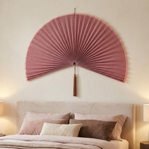 May include: A large, semi-circular wall fan in a dusty rose color with a pleated texture and a matching fabric tassel hanging from the center. The fan is mounted horizontally, displaying a uniform, radiating pattern of folds across its surface.