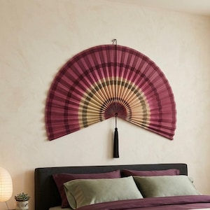 K&ouml;nnte beinhalten: Ein gro&szlig;er, halbkreisf&ouml;rmiger, an der Wand montierter Faltf&auml;cher mit einem Verlaufsmuster in tiefem Magenta, gedecktem Oliv und Cremet&ouml;nen. Der F&auml;cher besteht aus plissiertem Material und hat eine lange, dunkle Zierquaste, die in der Mitte h&auml;ngt.