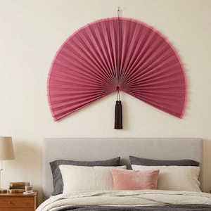 May include: A large, semi-circular wall fan in a deep magenta color with a pleated texture and a fringed edge. The fan is mounted on the wall and features a dark brown decorative tassel hanging from the center point of the fan's base.