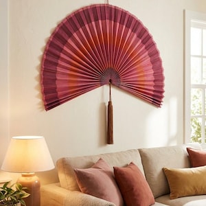 Op de afbeelding: Een grote, waaiervormige wanddecoratie in roze en oranje tinten hangt aan een crèmekleurige muur. Een bruine kwast hangt in het midden. De waaier hangt boven een beige bank met roze en roestkleurige kussens.
