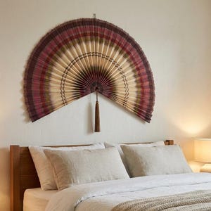 May include: A large, semi-circular wall fan featuring a pleated design with concentric bands of deep burgundy, muted olive green, and cream. The fan is finished with a dark brown decorative tassel hanging from the center point of the base.
