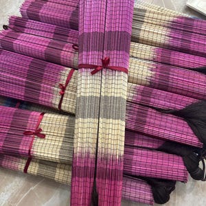 K&ouml;nnte beinhalten: Eine Sammlung rechteckiger Bambusmatten, die in einem Stapel angeordnet sind. Jede Matte weist ein Verlaufsmuster aus horizontalen Streifen in Magenta, Dunkellila und Cremefarben auf. Mehrere Matten sind mit d&uuml;nnen roten B&auml;ndern geb&uuml;ndelt.
