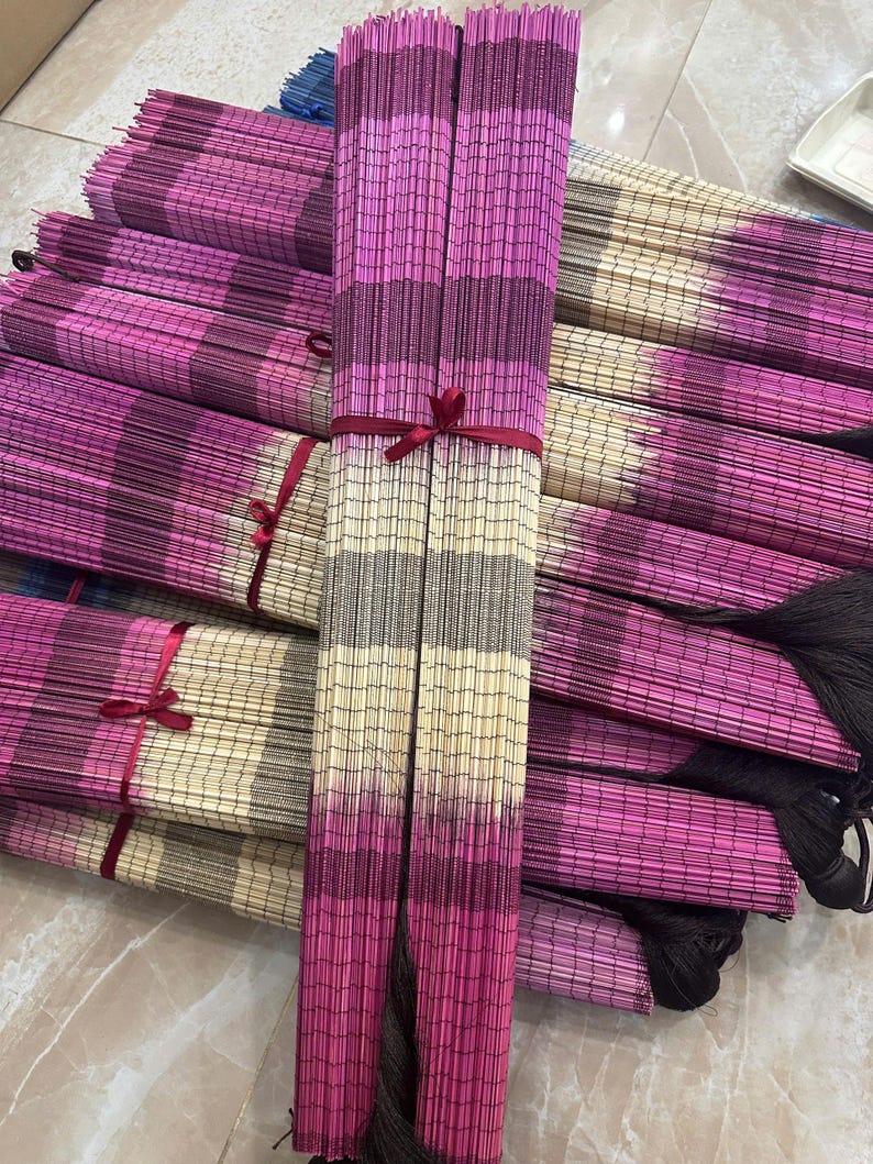 K&ouml;nnte beinhalten: Eine Sammlung rechteckiger Bambusmatten, die in einem Stapel angeordnet sind. Jede Matte weist ein Verlaufsmuster aus horizontalen Streifen in Magenta, Dunkellila und Cremefarben auf. Mehrere Matten sind mit d&uuml;nnen roten B&auml;ndern geb&uuml;ndelt.