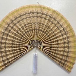 K&ouml;nnte beinhalten: Ein gro&szlig;er, halbkreisf&ouml;rmiger Wandf&auml;cher mit einem plissierten Design aus abwechselnden Streifen in hellem Beige, Sandfarben und zartem Gr&uuml;n. Der F&auml;cher ist in der Mitte mit einem kleinen Metallring befestigt und hat eine lange, wei&szlig;e Quaste.