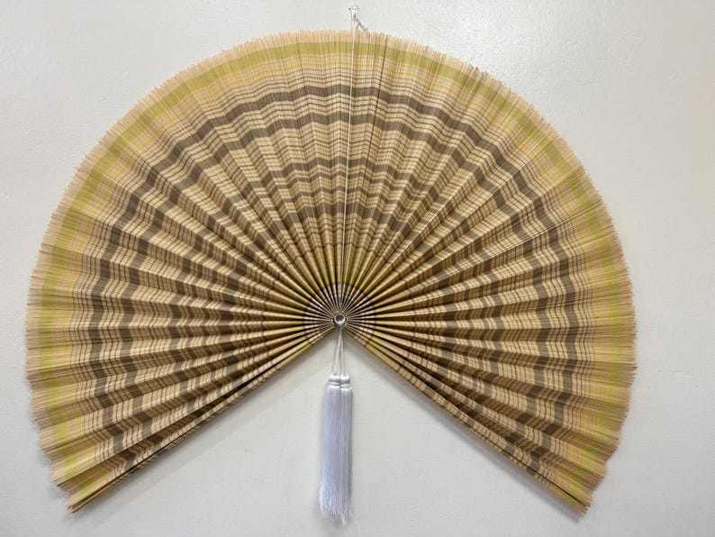 K&ouml;nnte beinhalten: Ein gro&szlig;er, halbkreisf&ouml;rmiger Wandf&auml;cher mit einem plissierten Design aus abwechselnden Streifen in hellem Beige, Sandfarben und zartem Gr&uuml;n. Der F&auml;cher ist in der Mitte mit einem kleinen Metallring befestigt und hat eine lange, wei&szlig;e Quaste.