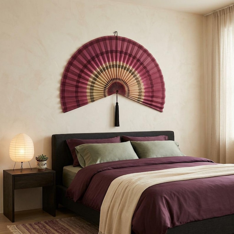 K&ouml;nnte beinhalten: Ein gro&szlig;er, halbkreisf&ouml;rmiger, an der Wand montierter Faltf&auml;cher mit einem Verlaufsmuster in tiefem Magenta, gedecktem Oliv und Cremet&ouml;nen. Der F&auml;cher besteht aus plissiertem Material und hat eine lange, dunkle Zierquaste, die in der Mitte h&auml;ngt.