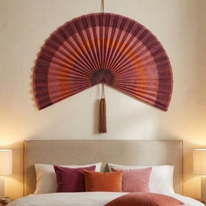 Könnte beinhalten: Ein großer, halbkreisförmiger Wandfächer mit einem plissierten Design in den Farben Dunkelburgunder, Magenta und gebranntem Orange. Der Fächer ist oben aufgehängt und hat eine lange, passende braune Quaste, die am Mittelpunkt hängt.