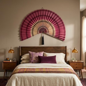 K&ouml;nnte beinhalten: Ein gro&szlig;er, halbkreisf&ouml;rmiger Wandf&auml;cher mit plissiertem Design in Magenta, Dunkelrosa und Creme, akzentuiert durch eine dunkelblaue Quaste, die in der Mitte h&auml;ngt. Der F&auml;cher ist an einer Wand &uuml;ber einem Holzkopfteil und Bett montiert.