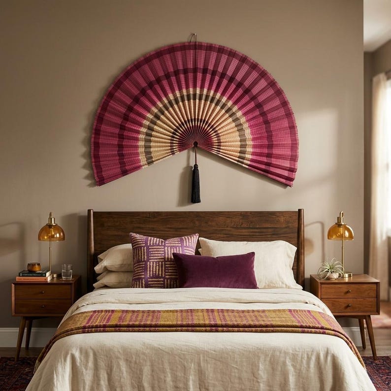 K&ouml;nnte beinhalten: Ein gro&szlig;er, halbkreisf&ouml;rmiger Wandf&auml;cher mit plissiertem Design in Magenta, Dunkelrosa und Creme, akzentuiert durch eine dunkelblaue Quaste, die in der Mitte h&auml;ngt. Der F&auml;cher ist an einer Wand &uuml;ber einem Holzkopfteil und Bett montiert.