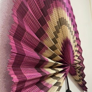 K&ouml;nnte beinhalten: Ein gro&szlig;er, halbkreisf&ouml;rmiger Wandf&auml;cher aus d&uuml;nnen, geflochtenen Schilfrohren. Der F&auml;cher zeigt ein sich wiederholendes Zickzackmuster in den Farben leuchtendes Magenta, tiefes Violett und nat&uuml;rliches Beige. Eine schwarze Quaste h&auml;ngt am Drehpunkt.