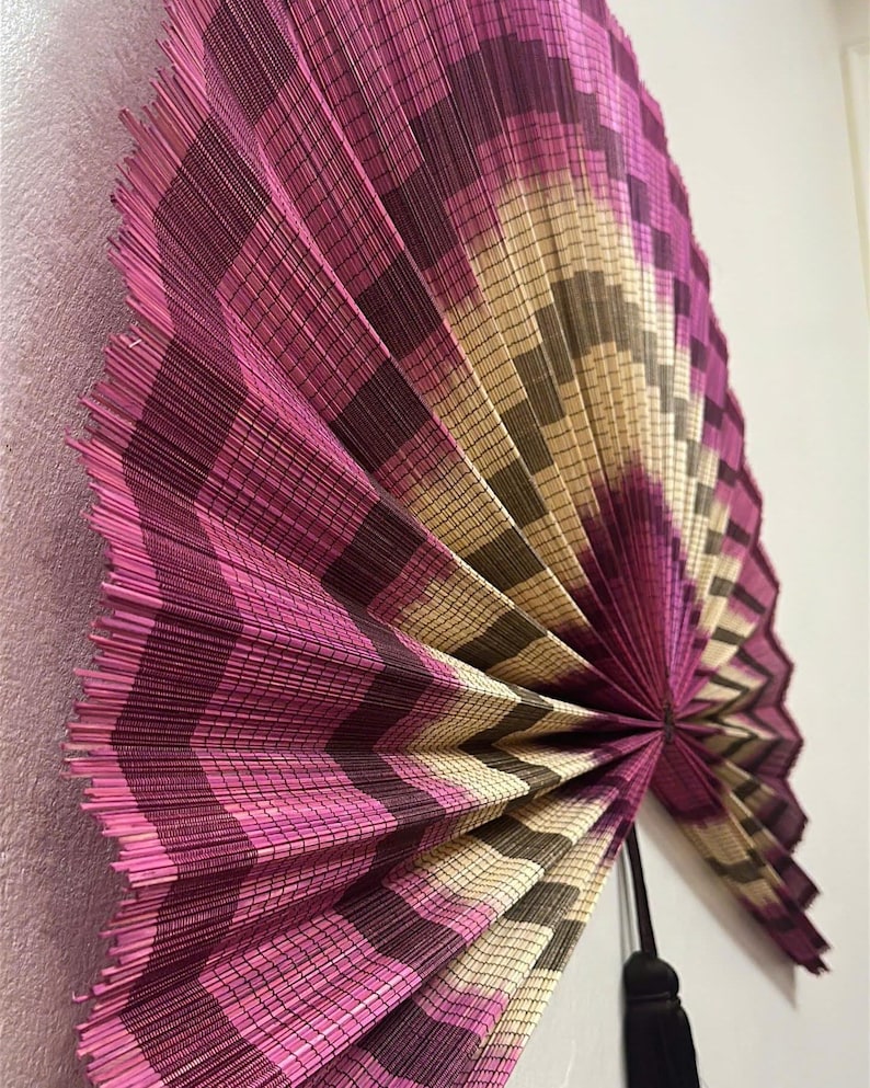 K&ouml;nnte beinhalten: Ein gro&szlig;er, halbkreisf&ouml;rmiger Wandf&auml;cher aus d&uuml;nnen, geflochtenen Schilfrohren. Der F&auml;cher zeigt ein sich wiederholendes Zickzackmuster in den Farben leuchtendes Magenta, tiefes Violett und nat&uuml;rliches Beige. Eine schwarze Quaste h&auml;ngt am Drehpunkt.