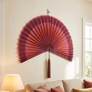 Puede incluir: Un gran abanico de pared decorativo en tonos rosa y burdeos, con efecto degradado. El abanico está montado en una pared y tiene una borla marrón colgando de la parte inferior. Un artículo de decoración del hogar único.