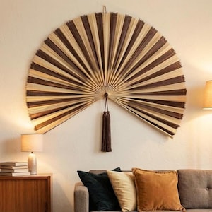 Può includere: Un grande ventaglio da parete semicircolare realizzato con fibre naturali che alternano il beige chiaro al marrone scuro. Il ventaglio presenta un design plissettato che si irradia da un punto centrale, con una lunga nappa marrone.