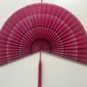 K&ouml;nnte beinhalten: Ein gro&szlig;er, halbkreisf&ouml;rmiger Faltf&auml;cher in Magenta- und Dunkelrosat&ouml;nen. Der F&auml;cher hat ein plissiertes Design mit horizontalen Streifen in verschiedenen Rosat&ouml;nen und einen zentralen Drehpunkt. Eine lange, d&uuml;nne rote Quaste h&auml;ngt unten mittig.