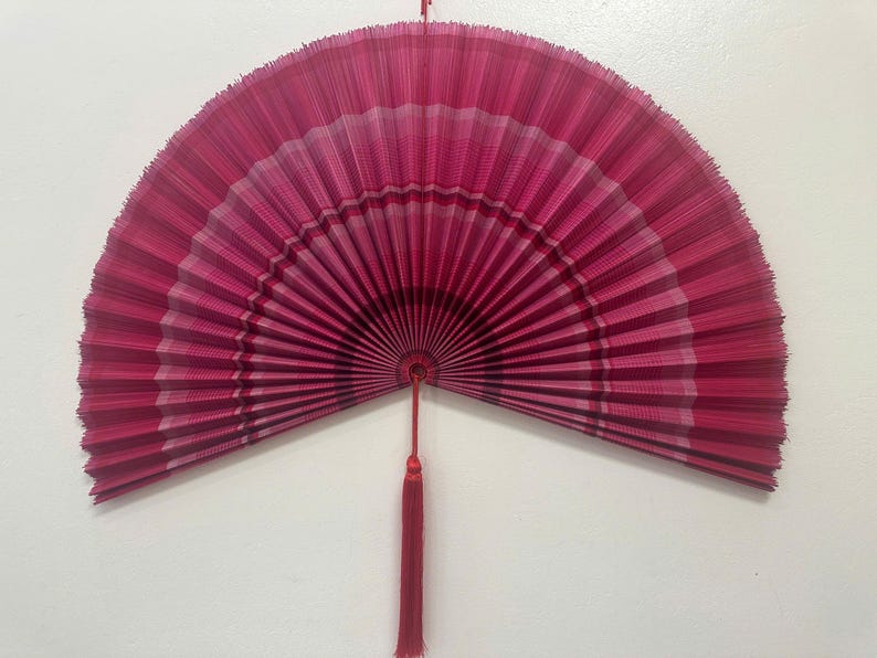 K&ouml;nnte beinhalten: Ein gro&szlig;er, halbkreisf&ouml;rmiger Faltf&auml;cher in Magenta- und Dunkelrosat&ouml;nen. Der F&auml;cher hat ein plissiertes Design mit horizontalen Streifen in verschiedenen Rosat&ouml;nen und einen zentralen Drehpunkt. Eine lange, d&uuml;nne rote Quaste h&auml;ngt unten mittig.