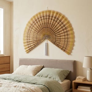 K&ouml;nnte beinhalten: Ein gro&szlig;er, halbkreisf&ouml;rmiger Wandf&auml;cher mit plissiertem Design und einem Karomuster in Beige-, Braun- und ged&auml;mpften Gelbt&ouml;nen. Der F&auml;cher besteht aus d&uuml;nnen, strahlenf&ouml;rmig angeordneten Lamellen und hat eine hellgraue Quaste in der Mitte.