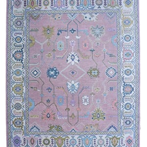 Könnte beinhalten: Ein rechteckiger Teppich mit floralem Muster in Rosa, Blau, Grün und Weiß. Der Teppich hat ein zentrales Design und einen Rand aus sich wiederholenden geometrischen und floralen Motiven. Die Farbpalette umfasst sanfte Pastelltöne und gedämpfte Farben.
