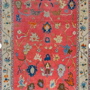 Könnte beinhalten: Ein rechteckiger Teppich mit einem leuchtend roten Mittelfeld. Der Teppich hat einen cremefarbenen Rand mit floralen und geometrischen Mustern in Blau, Braun und Orange. Das Mittelfeld ist mit stilisierten Blumenmotiven in verschiedenen Farben verziert.
