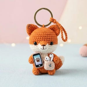 May include: A crocheted fox keychain featuring orange and white yarn, black safety eyes, and a stitched nose. The fox holds a small black smartphone with a paw print screen and a beige pouch containing white wireless earbuds, attached to a metal ring.