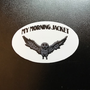 May include: White oval sticker with the text "MY MORNING JACKET" in black lettering. A black and white illustration of an owl with its wings spread is centered on the sticker.