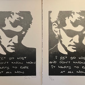 May include: A black and white print of a man's face with the text "I get so lost and don't know how it hurts to care at all now". The print is numbered 1/20 and signed "Saule 2024".