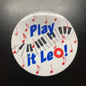 May include: White round sticker with a black and white piano keyboard design. The sticker says "Play it Leo!" in blue letters and has red musical notes around the edge.