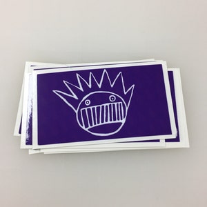 May include: A stack of purple stickers with a white cartoon face with a crown of spikes and a mouth with teeth.