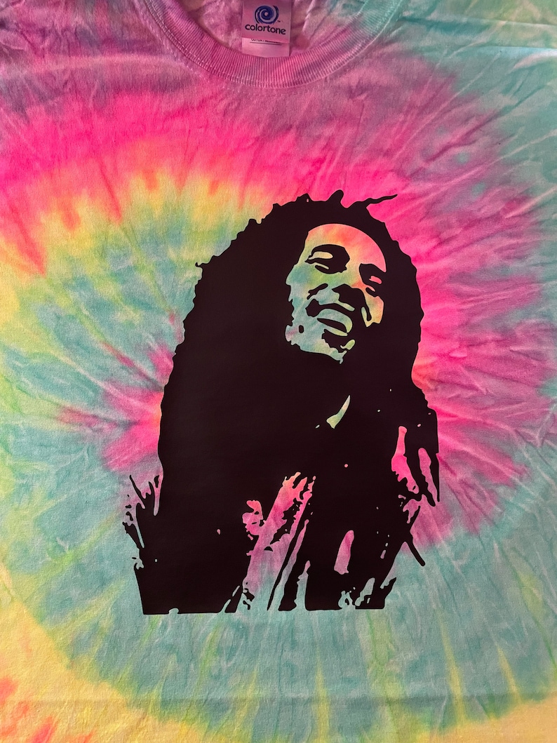 May include: A tie-dye t-shirt featuring a black silhouette of Bob Marley on the front. The shirt is a light blue, yellow, pink, and green colour.