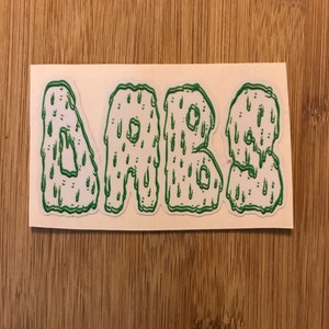 May include: A white sticker with the word "DABBS" in green, dripping letters. The letters have a cartoonish, melted appearance, with green drips. The sticker is rectangular and placed on a wooden surface.