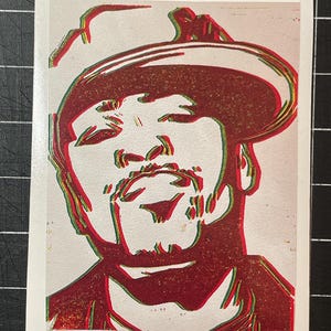 May include: A red, green, and white linocut print of a person wearing a hat. The portrait has a vintage, slightly distressed aesthetic. The image is on a white background.