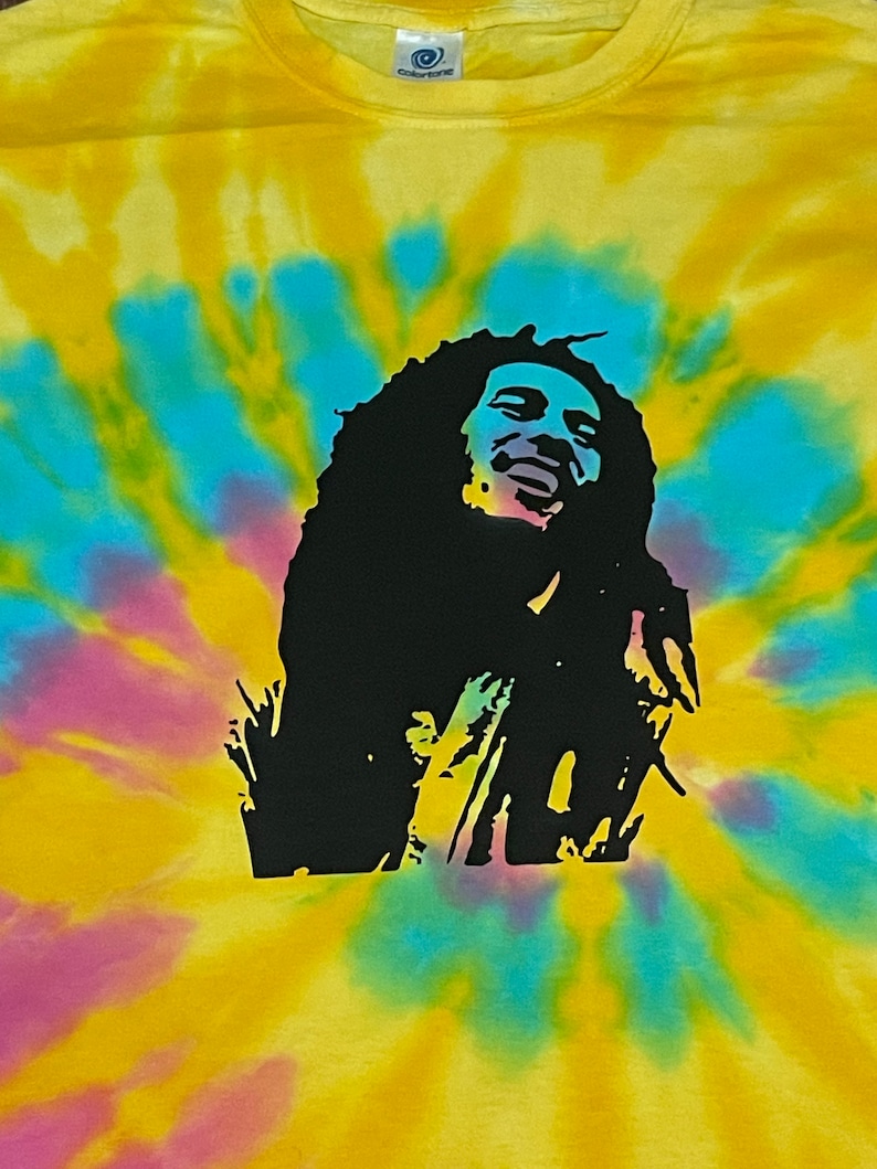 May include: A yellow, blue and pink tie-dye t-shirt featuring a black silhouette of Bob Marley singing.