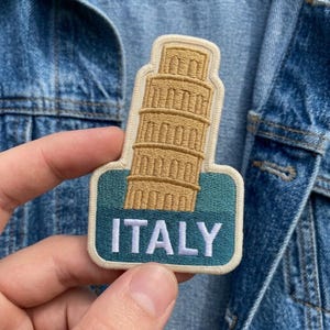 May include: An embroidered patch featuring a tan Leaning Tower of Pisa graphic set against a teal and dark blue background with the word ITALY stitched in white capital letters, finished with a cream-colored border and rounded edges.