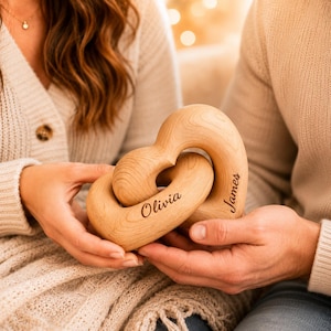 May include: A light brown wooden sculpture featuring two interlocking heart shapes. The surface of the wood displays a natural grain pattern. The name Olivia is engraved on one heart, and the name James is engraved on the other heart.