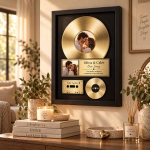 Könnte beinhalten: Ein schwarzer Wandrahmen mit einer großen goldenen Schallplatte mit einem Paarfoto in der Mitte, einer rechteckigen Plakette mit dem Text Olivia & Caleb Our Song, einer goldenen Kassette und einer kleinen goldenen Platte mit Herzsymbol.