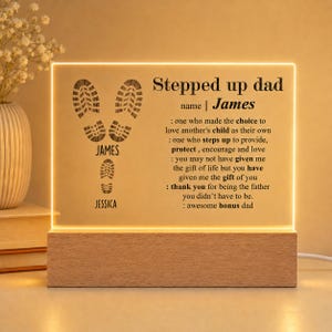 May include: A rectangular acrylic plaque with warm LED backlighting set in a light wood base. The plaque features black text reading Stepped up dad, James, and a poem about fatherhood, alongside graphic footprints labeled James and Jessica.