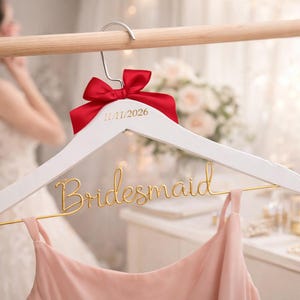 Könnte beinhalten: Ein weißer Holzkleiderbügel mit roter Schleife und goldenem Text mit der Aufschrift "11/11/2026" und "Brautjungfer". Ein rosa Kleid hängt an dem goldenen Draht. Der Hintergrund ist verschwommen, mit einer Braut im Brautkleid.