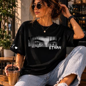 Könnte beinhalten: Schwarzes T-Shirt mit einer schwarz-weißen Grafik von Augen und dem Namen "ETHAN" in Weiß. Das T-Shirt wird mit hellblauen Jeans getragen. Ein kleines "E" und ein Herz befinden sich auf dem linken Ärmel.