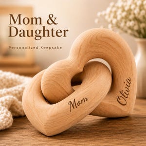 May include: Two interlocking light-colored wooden heart shapes with smooth, rounded edges. The left heart is engraved with the word Mom, and the right heart is engraved with the name Olivia. The wood grain is visible throughout the surface of both hearts.