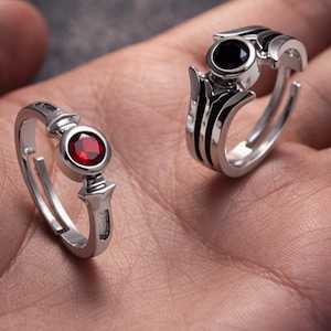 May include: Two silver rings with ornate designs. One ring features a round, red gemstone, while the other has a black gemstone. The rings are displayed on a hand, showcasing their intricate details and craftsmanship.