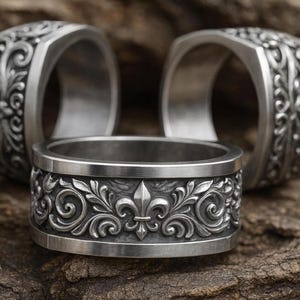 May include: Three silver rings with ornate floral and fleur-de-lis designs. The rings are displayed on a textured, brown surface. The rings have a width of approximately 0.5 inches.