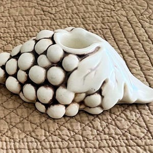 Pode incluir: Dois castiçais de cerâmica branca suja em forma de cachos de uvas com folhas. Os castiçais têm uma cor de destaque marrom escuro e são colocados em um fundo neutro. Castiçais decorativos.