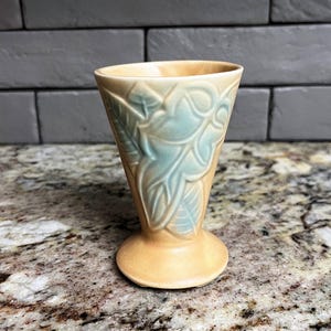 May include: A vintage, small vase with a flared top and a round base. The vase is a light tan color with a teal leaf design. The vase is approximately 5 inches tall.