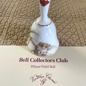 May include: A white porcelain bell with a gold rim and handle, featuring a red poppy and wheat field design. The bell has a small, detailed illustration of a hamster. Text on the card reads "Bell Collectors Club" and "Wheat Field Bell".