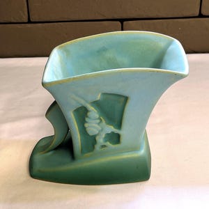 May include: A vintage art deco vase in shades of teal and green. The ceramic vase features a stylized design with a geometric shape and a decorative element. The vase has a unique, asymmetrical form.