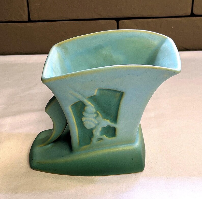 May include: A vintage art deco vase in shades of teal and green. The ceramic vase features a stylized design with a geometric shape and a decorative element. The vase has a unique, asymmetrical form.