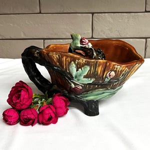 May include: A decorative ceramic planter with a brown interior and a dark handle. The exterior features a tree bark design with green leaves and red berries. A cluster of vibrant pink roses rests beside the planter.