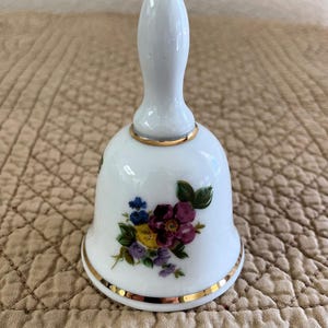 May include: White ceramic bell with a gold rim and handle. The bell features a floral design with purple, blue, yellow, and green flowers. The bell is a decorative item.