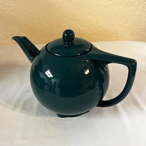 Op de afbeelding: Een teal-kleurige keramische theepot met een afgeronde body, een gebogen handvat en een tuit. Het deksel heeft een kleine knop bovenop. De theepot is glanzend en reflecteert licht.