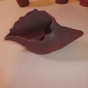 May include: A burgundy-colored ceramic decorative bowl with a unique, organic shape. The bowl has a wide, open mouth and a textured surface. The design resembles a seashell or a natural form, adding an artistic touch to any space.